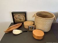 Wide shot of entire collection showing six items arranged on table against light grey wall: framed woven basket art, wooden canoe bowl, small round woven basket, bark spoon with carved handle, square decorated box, and oval wooden box.