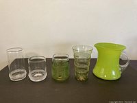 Front view of four art glasses and one art glass pitcher arranged side by side on dark surface against white background showing various shapes and surface designs