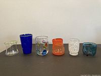 Full view of six art glasses in varied colors and styles on surface against white wall.