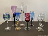 Full view of eight assorted tall art drinking glasses in varied colors and styles arranged on dark surface