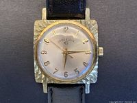 Close-up image of the Lord Elgin watch face showing textured gold-tone square case, silver dial with gold-tone numerals and hands, and black leather strap.