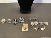 Full frontal view of all six pairs of cufflinks and one money clip arranged in two rows on black surface.