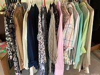 Photo shows several women's blouses hanging on a rack, featuring various colors and patterns including floral, plaid, and solids with long sleeves and collars.