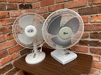 Two white table top fans side by side on a wooden table against a brick wall background. Both fans have metal grilles and bases with control buttons.