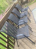 Four black metal frame patio chairs with mesh seats and backs arranged on a wooden deck against a railing. Chairs have curved armrests and are showing signs of outdoor wear but are in good condition.