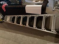 Aluminum extension ladder laid flat on the floor in a residential setting, showing full length with rectangular rungs and side rails.