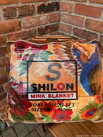 Front view of packaged blanket in plastic case, showing colorful bird and floral pattern and brand logo 'Shilon Mink Blanket'.