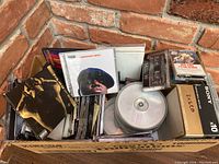 Photo of a cardboard box containing a mix of CDs and cassette tapes. Visible CD covers include a Greatest Hits album and other various titles. Cassette tapes are mixed with some in labeled boxes and some in clear plastic cases.