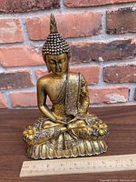 Seated Buddha statue with detailed hair curls and ornate gold-toned robe decorated with amber colored stones, placed on wooden surface with brick wall background, shows front and angled view of the statue.