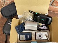 Top-down open box view showing Kodak Cameo film camera, Canon PowerShot SD300 digital camera with battery and charger, and Sony Cyber-shot DSC-T7 digital camera with charging accessories.