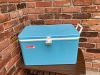 Blue Coleman cooler placed on a wooden top against brick walls showing the front, white handle, and hinged lid.