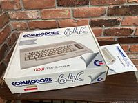 Closed original Commodore 64C box on wooden table with brick wall background, manual edge visible