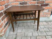 Mid century style wood veneer end table with tapered legs positioned outdoors against brick wall on stone pavers.