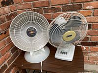 Image showing two tabletop appliances against a brick wall background, a white electric heater with a reflective surface and brand label 'Proheat 1500 Watt' on the left, and a white electric fan with brand label 'Super' on the right.
