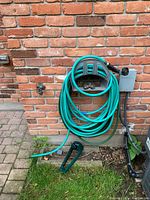 Photo shows garden hose wrapped around a metal reel holder mounted on a brick wall. Hose has nozzle attached and extends on ground with some slack.
