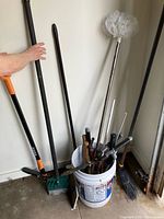 Wide view of garden tools including hedge trimmers, edger with orange handle, several long handled brooms, a white bucket holding various hand tools and shears.