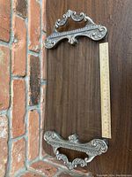 Top-down view of rectangular tray showing silver-plated ornate handles and frame with clear glass inside, placed on wood surface next to a ruler for scale.