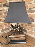 Full front view of the table lamp showing greyhound sculpture, book base and black shade placed on wooden surface against brick wall.