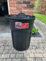 Black Rubbermaid 32 gallon garbage bin with lid shown outside on brick driveway near a brick wall and garage door.