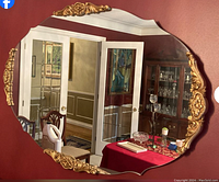 Full view of large oval mirror hanging on a burgundy wall, reflecting a dining area with glass cabinet and table.