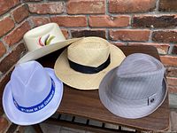 Three men's hats displayed on dark wooden table against brick wall: cowboy style hat, straw fedora, and gray pinstripe fedora.