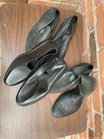 Photo shows five pairs of black Totes men's rubber overshoes arranged on a wooden surface near brick wall. Overshoes have an elastic panel on the side, designed to slip over regular shoes and protect from water or dirt. Size large fits men's approximate shoe size 9.5-10. Condition is lightly used with some creasing and signs of wear.