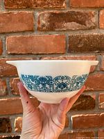 Photo showing side view of vintage Pyrex Horizon Blue Cinderella mixing bowl with teal blue patterned design against brick background held by a hand.