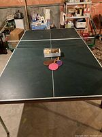 Full view of green table tennis table with paddles and balls on table surface in basement setting.