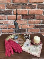 Full lot of vanity items arranged on table, showing gloves, bird statue, brass mirror tray, candle in holder, and glass ring dish.