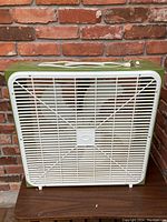 Vintage green and white box fan photographed against a brick wall, showing front protective grille and fan blades.