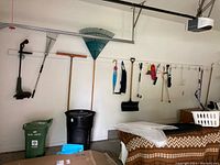 Photo of garage wall with garden tools hanging on a white hook rack, showing electric Black & Decker trimmer, several rakes, shovels, mop, and other tools.