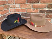 Two western style hats on a wooden surface against a brick background. One black felt hat with feather, one brown felt hat with feather and decorative band.