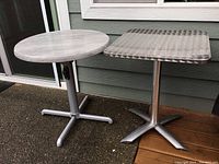 Two outdoor folding tables side by side, one round plastic top on gray metal base, one square stainless steel top on metal base