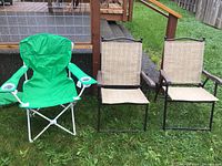 Photo of three folding chairs on grass: a green fabric camping chair with white metal legs and two beige sling mesh chairs with black metal frames and armrests.