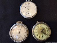 Three vintage pocket watches displayed together, showing different dial designs and metal cases.