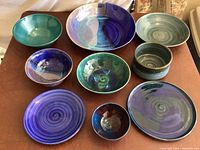 Photo showing nine pottery bowls in various shades of blue and green, arranged on a flat surface.