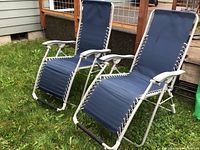 Two blue folding reclining chairs placed outdoors on grass. Both chairs reclined flat showing metal frames, fabric seats, cord lacing, and padded armrests.