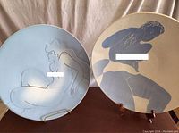 Two ceramic platters with blue and white powder finish nude art, displayed on stands against a beige fabric backdrop.