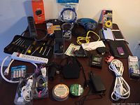 Overview of all the assorted electronic items displayed on a table including cables, adapters, chargers, surge protectors, headphones, small tools, an iPod, an old iPhone, and Apple TV device.