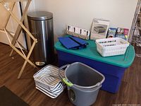 Full view of lot showing SimpleHuman stainless steel trash can, wooden drying rack, plastic storage tote, blue folding bag, white laundry basket, and striped fabric storage bin