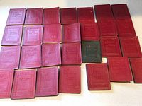 Set of 28 miniature leather-bound books arranged on a flat surface, 27 red covers and 1 black cover visible.