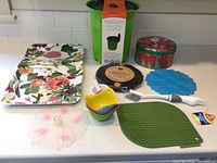 Full lot contents showing compost bin, trays, measuring cups, pot holder, trivet, tin container, cleaning brush, pink flower-shaped item