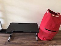 Red canvas travel bag and black lap desk side by side on floor near a wall.