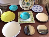Overview photo showing various ceramic plates and bowls of different styles including yellow, turquoise, square floral, and plain white plates and bowls arranged on a wooden surface.