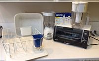 White Rubbermaid dish drainer, Proctor Silex blender, DeLonghi toaster oven, Brita water filter pitcher, and box of filters on kitchen counter.