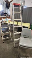 Tall white metal step ladder and smaller step stool beside two gray plastic stacking chairs in warehouse setting.