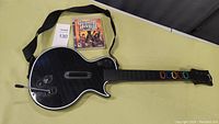 Black wireless Guitar Hero 3 guitar controller with strap and the game disc in case placed beside it.
