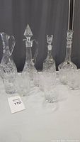 Six lead crystal decanters with varied stoppers and six matching whiskey glasses arranged on a white surface with dark backdrop.
