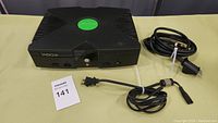 Front and top view of the black original Xbox console with green logo on top and two power/AV cables alongside.