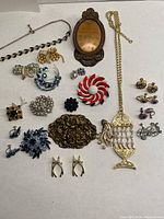 Photo showing an assortment of vintage costume jewelry pieces arranged on a flat surface including a large brass-tone brooch, colorful rhinestone brooches, multiple pairs of clip-on earrings, and a gold-tone necklace with pendant.